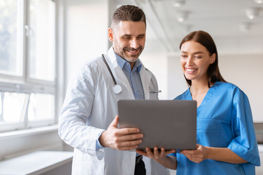 Happy Male Doctor And Female Nurse Talking, Working Together, Using Laptop, Discussing Patient Medical Checkup Results