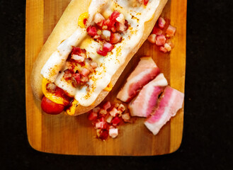 Homemade hot dog with bacon, melted cheese and mustard on wooden board and black background
