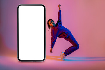 Active african american female hip-hop dancer dancing near huge smartphone with blank screen, neon background, mockup