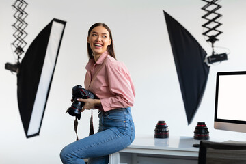Excited female photographer sitting on edge of table, holding her DSLR camera and smiling, photostudio interior