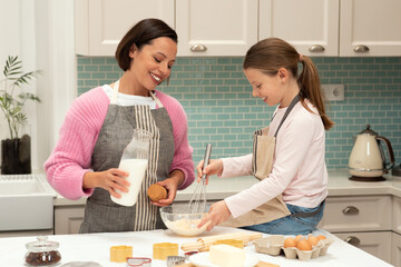 Smiling caucasian young mother and little daughter make cookie dough with milk in kitchen interior