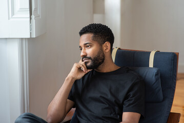 Young handsome black man sitting at home in the armchair and looking thoughtfully in the window