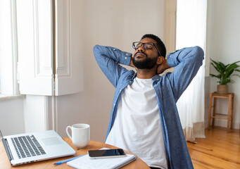 Tired young black man wearing glasses is stretching in front of his laptop at home office,...