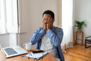 Tired young black man rubbing his face and irritated eyes with hands in front of his laptop at home...