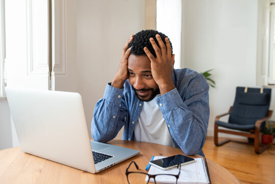Frustrated young African entrepreneur with sad grimace in front of his laptop at home office, noticed a big mistake he did, holding his face with hands, wearing blue shirt