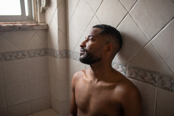 Young black shirtless sad man sitting alone in a bathroom. Sad, depressed, lonely, upset.