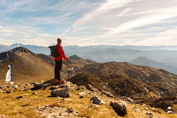 Obraz premium mountaineer man from behind with red clothes, backpack and trekking poles practicing high mountain hiking and contemplating the landscape. outdoor sport and adventure.
