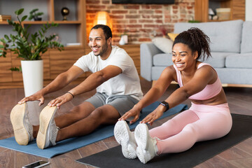 Glad young black couple in sportswear do leg stretching exercises on mat on floor, training together