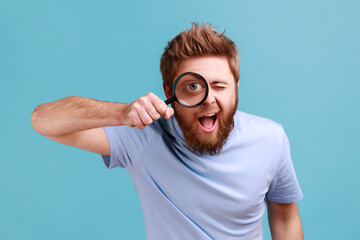 Man in blue T-shirt holding magnifying glass and looking at camera with big zoom eye, curious face.