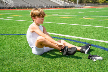 Male preteen youth football player sitting on field putting on cleats before practice