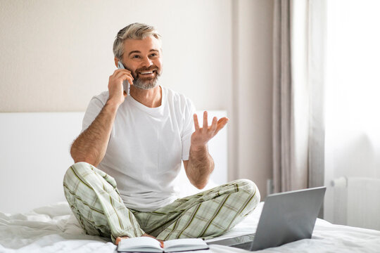 Happy man sitting on bed, using laptop, talking on phone