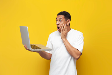 young shocked african american guy in white t-shirt uses laptop and shows surprise on yellow...