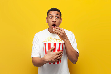 young shocked african american guy eating popcorn and watching movie on yellow isolated background,...