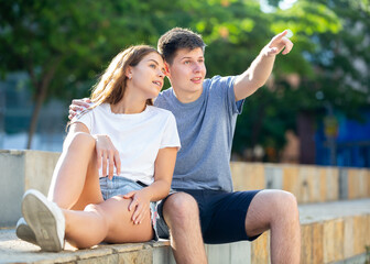 Young lady and young man are sitting on the step and pointing with fingers