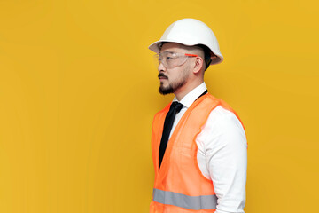 asian foreman in uniform stands sideways on yellow isolated background, korean civil engineer in hardhat and goggles