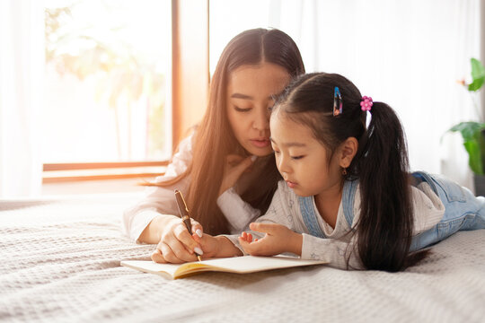 Asian Mother With Little Daughter Lie On Bed At Home And Learn To Write And Draw With Pen In Notebook