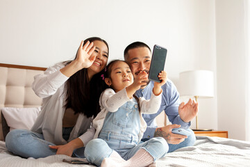 asian family sit on the bed and communicate via video call, little korean girl with mom and dad takes selfie
