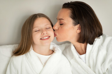 Fototapeta na wymiar Cheerful young lady kissing on cheek little caucasian girl in bathrobe, relaxing on white bed, enjoy spare time