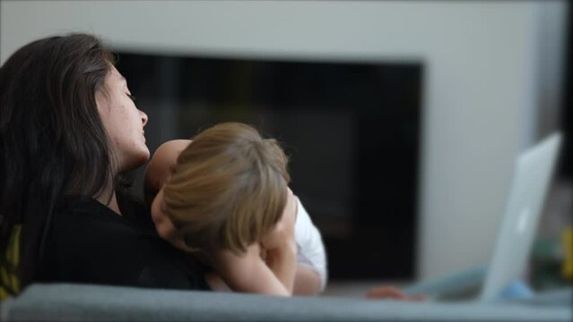 Mother trying to work and multi task with baby toddler at home sofa living room. Child bothering parent while working with laptop computer