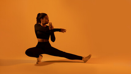 Fit black lady in sportswear stretching legs and arms, warming up, orange neon background, panorama, copy space