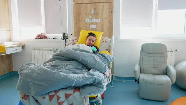 Tired But Happy New Mom Lies In Bed After Childbirth. Loving Mother Breastfeeding Her Baby In The Maternity Hospital Ward.