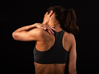 Female sporty muscular with ponytail doing stretching workout of the shoulders, blades in sport bra, holing the neck the hand on black studio background