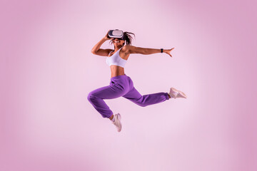 Fototapeta na wymiar Young black woman working out with 3D technology, jumping while wearing virtual reality headset, pink neon background