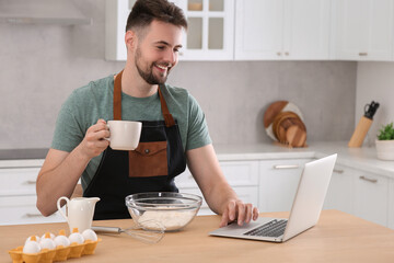 Happy man learning to cook with online video on laptop at table in kitchen. Time for hobby