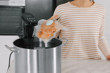 Woman putting vacuum packed meat into pot with sous vide cooker in kitchen, closeup. Thermal...