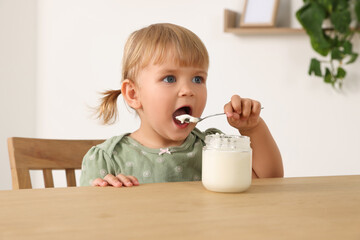 Cute little child eating tasty yogurt with spoon at wooden table indoors