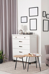 Empty frames hanging on white wall, chest of drawers and wooden tables indoors