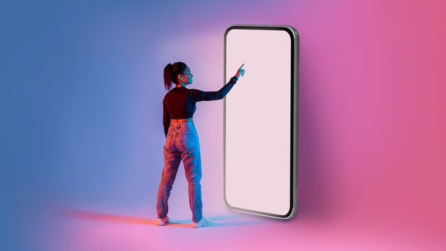 African american lady touching screen of giant smartphone in neon light, mockup for mobile app or website design
