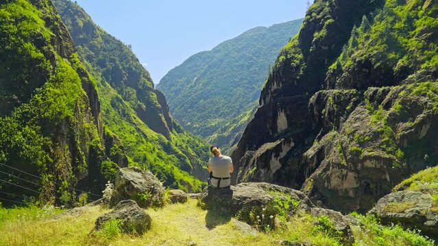 Moving towards the back of a woman sitting on the edge of a cliff overlooking a vast valley and mountainous landscape, Annapurna Circuit, Nepal