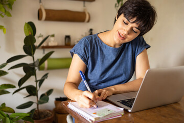 Young latin woman with a short hair taking notes from laptop at the home office. She is sitting in the apartment full of plants.