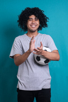 Smiling curly man holds soccer ball clapping hands, watching football match isolated on blue background