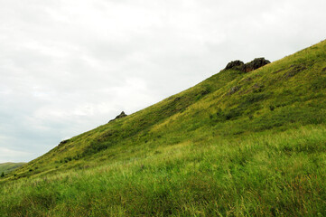 A steep slope of a high mountain overgrown with grass and rocky formations on top under a cloudy summer sky.