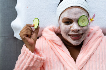 Portrait of happy plus size african american woman with beauty face mask and cucumber slices at spa