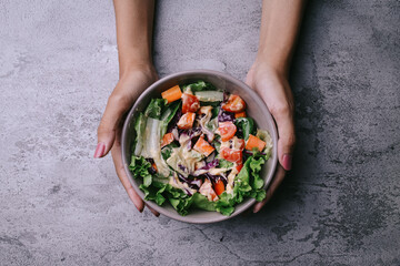Cropped shot of hand serving vegan salad from green leaves mix and vegetables. Top view on gray stone table.