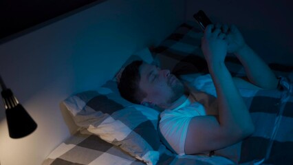 Man serious concentrated man chatting, typing message, looking at mobile mobile smart phone in bed, bedroom instead of sleeping deep at night. Social media addiction. Sleep disorder, sleep disturbance