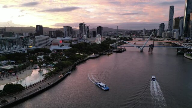 Drone shot of South Bank, shot over Brisbane River, camera orbits The Wheel of Brisbane. Citycat Boats crossing river. New Neville Bonner Bridge in construction in background, Sunset.