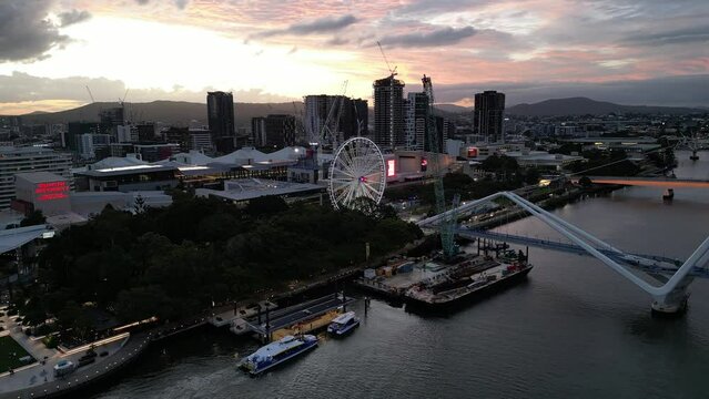 Drone shot of South Bank, shot over Brisbane River, camera tracking towards The Wheel of Brisbane. Citycat Boats at South Bank pontoon. New Neville Bonner Bridge in construction in background, Sunset.