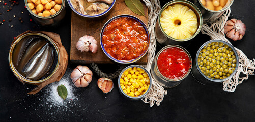 Canned vegetables, beans, fish and fruits in tin cans on black background. Food stocks.