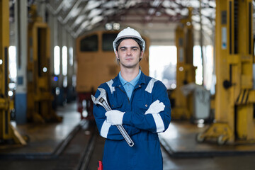 Male engineer worker working in industry factory, standing with crossed arms and holding industrial spanner, wearing safety uniform, helmet. Male technician worker at work in locomotive garage