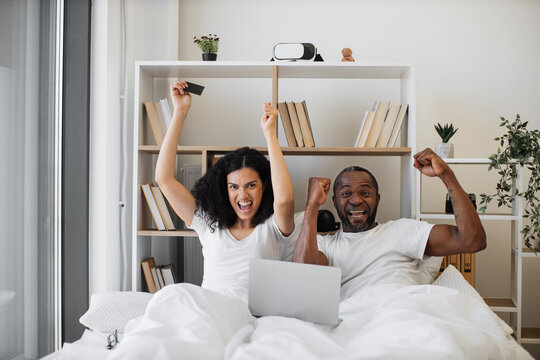 Emotional african american family of two raising hands in excitement while sitting in bed with laptop on blanket and credit card in hand. Thrilled married couple making successful deal online at home.