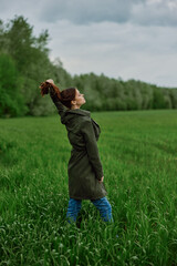 a beautiful woman in a dark coat stands in a green field in the spring in rainy weather and straightens her hair