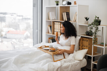 Beautiful young lady holding delicious croissant in hand while enjoying view out of panoramic window of white bedroom in morning. Pretty woman waiting for her husband to join for coffee in bed.
