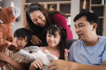 Cheerful and playful young Asian family with father along with mother and son and daughter sitting on couch and playing with rabbit stuff toy at home