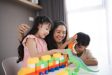Cute little Asian daughter focused while playing and learning with smiling and cheerful mother with toy train and assembling lego game blocks at home