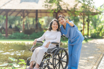 Young asian care helper with asia elderly woman on wheelchair relax together park outdoors to help and encourage and rest your mind with green nature. Pointing