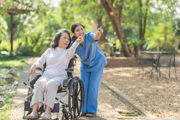 Young asian care helper with asia elderly woman on wheelchair relax together park outdoors to help and encourage and rest your mind with green nature. Pointing
